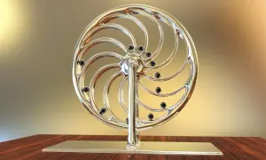 Glänzendes Rad mit schwarzen Kugeln, das an ein Perpetuum mobile erinnert, steht auf einem Holztisch.